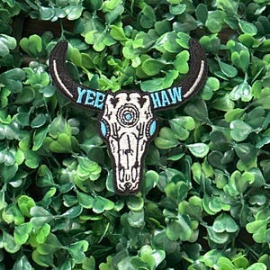 May include: Embroidered patch featuring a white bull skull with black horns and accents. The text "YEE HAW" is embroidered in blue across the top. The patch is set against a backdrop of green foliage.