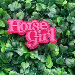 Puede incluir: Un parche bordado rosa con las palabras "Horse Girl" en una fuente cursiva. El parche está sobre un fondo de follaje verde.