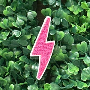 Könnte beinhalten: Ein pinkfarbener Blitz-Aufnäher mit weißem Rand. Der Aufnäher ist mit einem strukturierten Design bestickt. Der Hintergrund ist eine dichte Anordnung grüner Blätter, die einen kontrastierenden Hintergrund bilden.