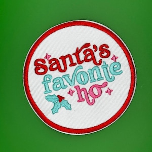 Peut inclure: Écusson brodé circulaire avec le texte "Santa's Favorite Ho" en rouge, turquoise et rose. L'écusson a un fond blanc et une bordure rouge. Une branche de houx est également présente.