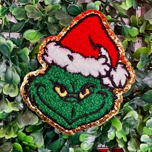 May include: A green Grinch face with a red Santa hat, white trim, and gold glitter edging. The Grinch's face has yellow eyes and a grumpy expression. The image is a Christmas-themed decoration.
