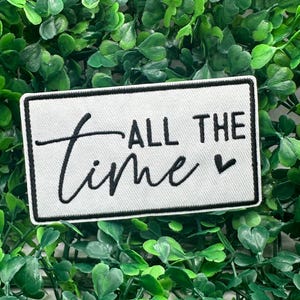 Puede incluir: Un parche rectangular blanco con un borde negro y las palabras "ALL THE time" en una fuente manuscrita. Un pequeño corazón negro está al lado de la palabra "time". El parche está sobre un fondo de hojas verdes.
