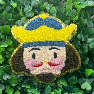 Puede incluir: Un parche bordado colorido que representa un personaje de dibujos animados con un sombrero amarillo, detalles azules y un borde dorado brillante. El personaje tiene la piel clara, mejillas rosadas y un bigote rojo. El parche está sobre un fondo de follaje verde.