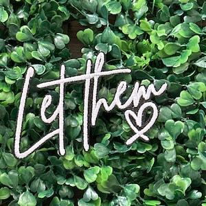 May include: A white embroidered patch with the words "Let them" in a cursive font, and a heart symbol. The patch has a black border and is set against a background of green leaves.