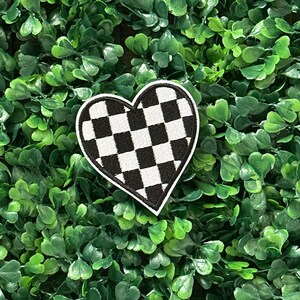 May include: A heart-shaped patch featuring a black and white chequered design. The patch is edged in white and is set against a background of green leaves.