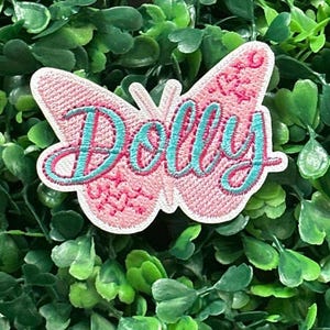 May include: A pink butterfly-shaped embroidered patch with the name "Dolly" in teal script. The patch has a white border and is set against a background of green foliage.