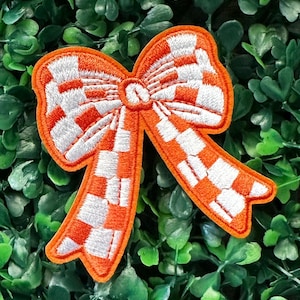 Puede incluir: Parche de lazo naranja y blanco a cuadros. El lazo está bordado con hilo blanco sobre una tela naranja.