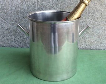 May include: A silver metal ice bucket with two handles, holding a bottle of champagne.