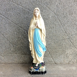 May include: A white statue of the Virgin Mary, standing on a base with a blue and white robe. The statue is in a praying position with her hands clasped together.