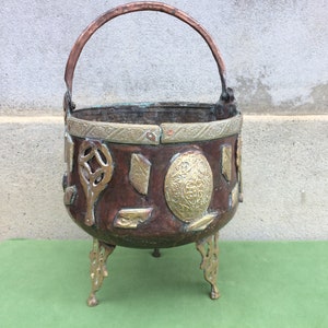 May include: A copper and brass cauldron with a handle and three legs. The cauldron is decorated with intricate brass designs.