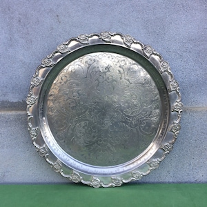 Può includere: Un vassoio da portata in argento con un design ornato e in rilievo. Il vassoio ha un bordo a festoni e una forma circolare.