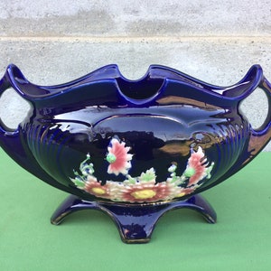 May include: A cobalt blue ceramic planter with two handles and a three-legged base. The planter is decorated with raised floral designs in shades of pink, yellow, and green.