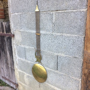 May include: A brass pendulum with a lyre-shaped design and a round weight hanging from a brick wall.