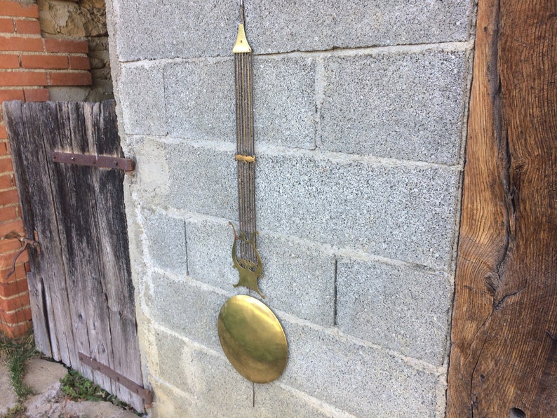 May include: A brass pendulum with a lyre-shaped design and a round weight hanging from a brick wall.