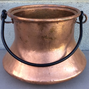 May include: A copper pot with a black iron handle. The pot has a wide base and a narrow top, and it is hammered to give it a textured surface.