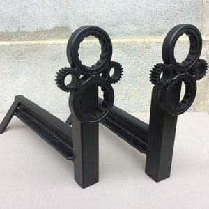 May include: A pair of black metal fireplace andirons with a gear design. The andirons are made of heavy-duty metal and have a sturdy construction.