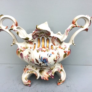 Puede incluir: Un tazón de porcelana blanca con dos asas y un diseño floral en tonos rosa, naranja y azul. El tazón tiene un borde festoneado y se asienta sobre una base de tres patas.