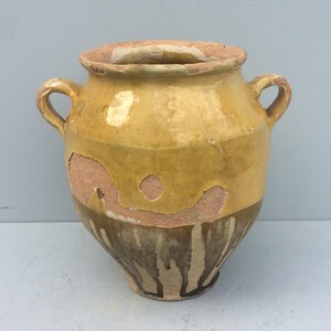 以下が含まれることがあります： 黄色と茶色のセラミック製の壺で、2つの取っ手が付いています。壺は幅が広く丸みを帯びたボディと、わずかにフレアしたリムを持っています。表面には摩耗や損傷が見られ、下の素材が露出している部分があります。壺はおそらくアンティークまたはヴィンテージ品です。