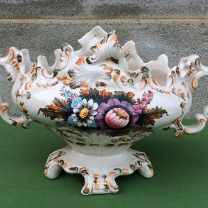 Może przedstawiać: Biała ceramiczna doniczka z ozdobnymi detalami i kwiatowymi wzorami w odcieniach różowego, fioletowego, niebieskiego, zielonego i pomarańczowego. Doniczka ma dwa uchwyty i trójnożną podstawę.