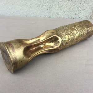 May include: A gold-colored metal vase with an intricate floral design. The vase is made from a repurposed artillery shell.