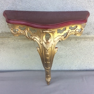 May include: A gold-painted wall shelf with ornate scrollwork and a burgundy-colored top.