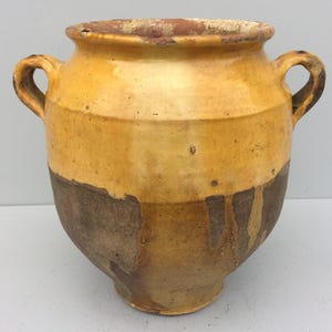 May include: A large, antique ceramic pot with two handles. The pot has a two-tone design, with a light yellow upper section and a dark brown lower section. The rim is a darker brown colour. The pot has a rustic, aged appearance.