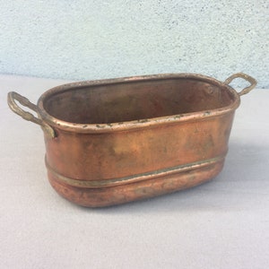 Könnte beinhalten: Ein ovaler Kupfertopf mit zwei Messinggriffen. Der Topf hat eine abgenutzte, gealterte Optik und ist perfekt, um Ihrem Zuhause einen rustikalen Touch zu verleihen.
