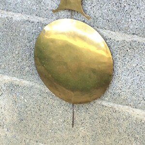 May include: A round, brass pendulum weight for a clock. The weight is hanging from a metal rod and is attached to a wall.