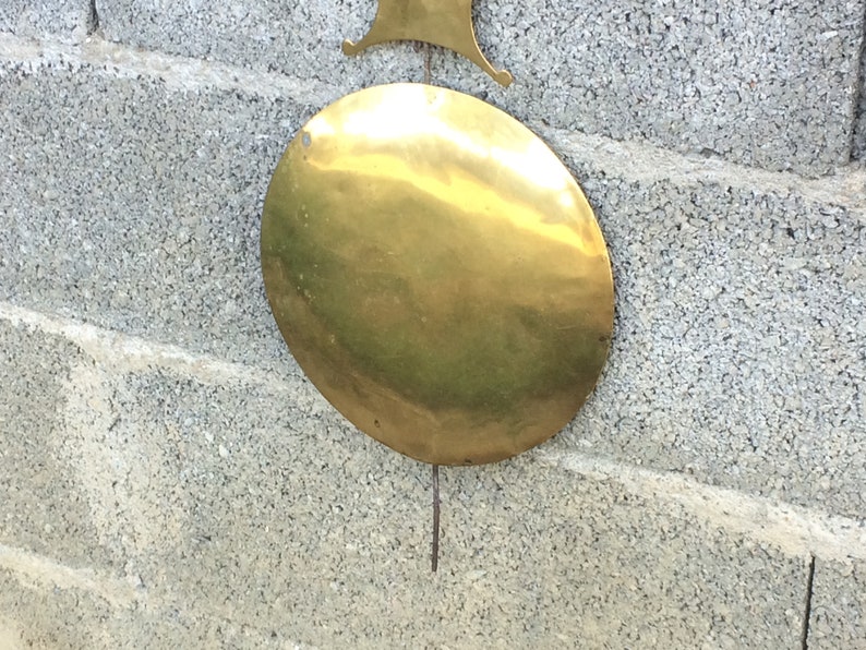 May include: A round, brass pendulum weight for a clock. The weight is hanging from a metal rod and is attached to a wall.