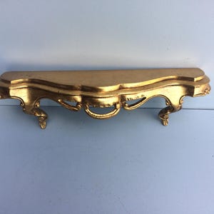 Pode incluir: Uma prateleira de madeira pintada de ouro com detalhes ornamentados e pés em forma de rolo. A prateleira é projetada para ser montada em uma parede.