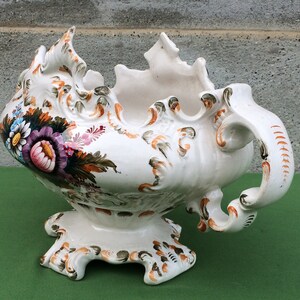 Pode incluir: Um vaso de cerâmica branco com um design floral. O vaso tem uma borda ondulada e uma alça. O vaso é decorado com flores em tons de rosa, roxo e laranja.