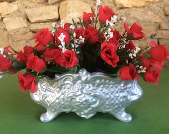 Op de afbeelding: Een zilveren plantenbak met een sierlijk ontwerp bevat een boeket rode rozen. De plantenbak heeft drie poten en is versierd met een reliëfpatroon.