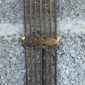May include: Close-up of a brass decorative element with a stylized design, attached to a series of vertical metal rods against a rough concrete wall.