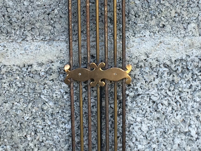 May include: Close-up of a brass decorative element with a stylized design, attached to a series of vertical metal rods against a rough concrete wall.