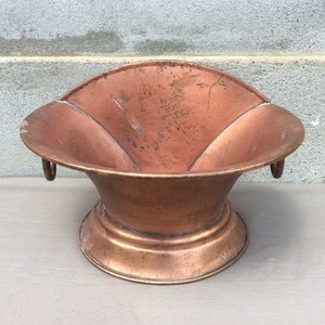 May include: A vintage copper planter with a wide, flared opening and two handles. The planter has a smooth, aged finish and a slightly rounded base.