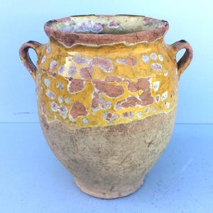 以下が含まれることがあります： 2つの取っ手が付いた、大きなアンティークのセラミック花瓶。花瓶は、黄褐色系の色合いで、使い古されたような外観をしています。上部の縁は濃い茶色で、本体は質感のある土のような外観をしています。