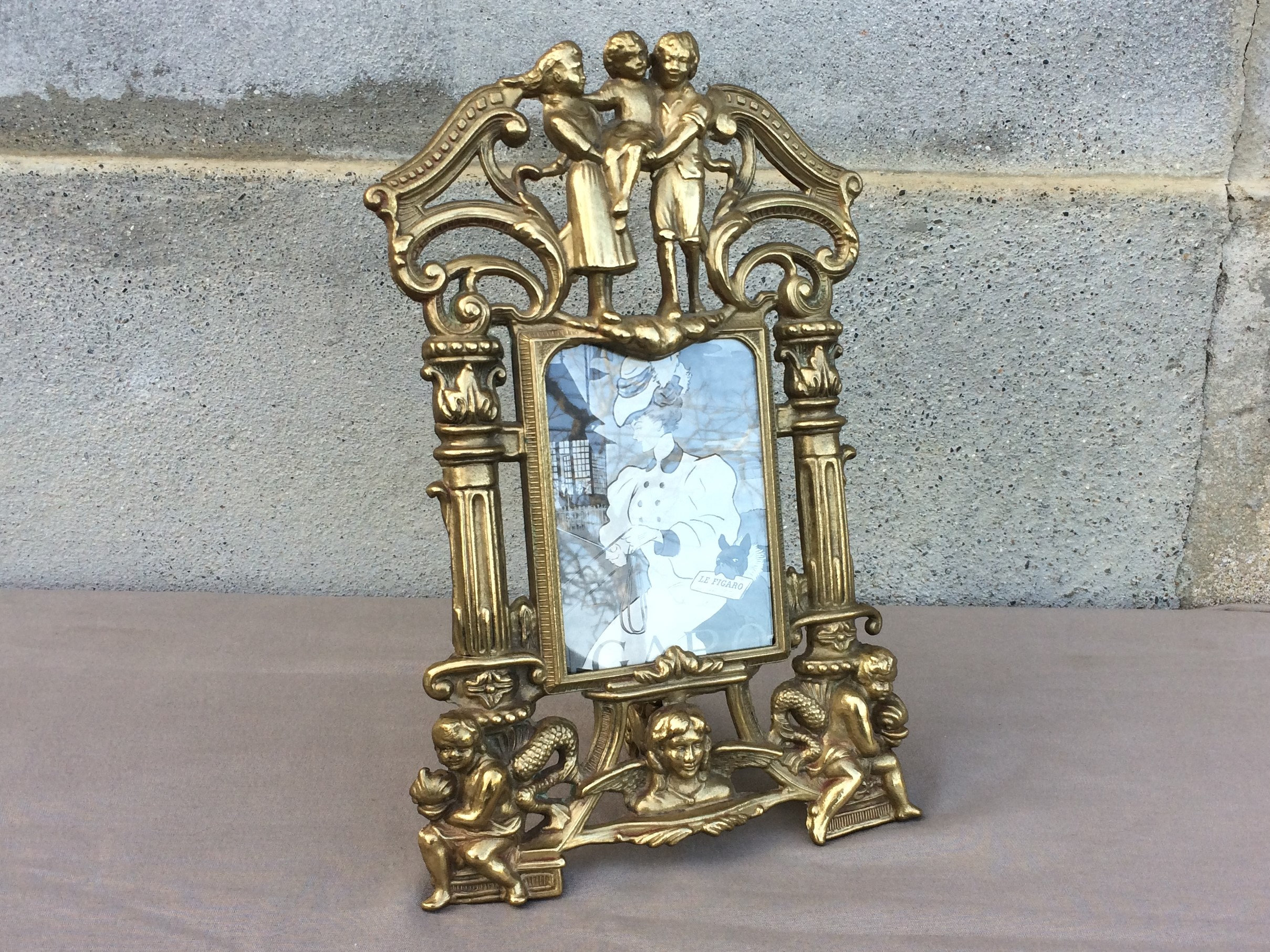 Puede incluir: Un marco de fotos de metal dorado con detalles ornamentados. El marco presenta figuras de querubines y una abertura rectangular central para una foto. El marco está diseñado para colocarse de pie.