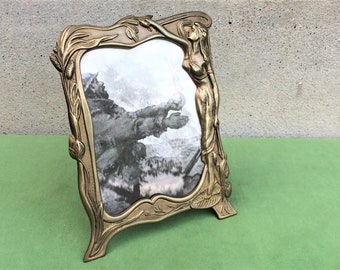 May include: A gold-toned metal picture frame with an ornate design featuring a woman figure and floral motifs. The frame has a rectangular opening for a photo and stands on a base.