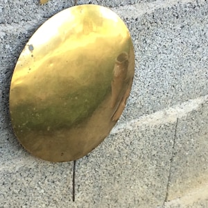 May include: A round, gold-colored metal disc hanging from a thin wire against a gray concrete wall.