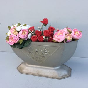 Könnte beinhalten: Ein silberner Blumentopf mit einem dekorativen Blumenmuster, gefüllt mit künstlichen rosa und roten Rosen.