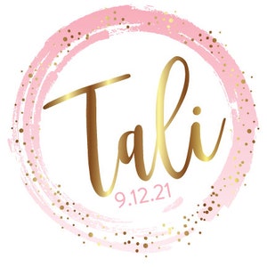 May include: A pink and gold circle with the name "Tali" written in gold script and the date "9.12.21" below it.