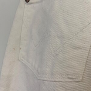 Op de afbeelding: Een close-up van een wit paar jeans met een achterzak. De zak heeft een gestikt ontwerp en een kleine knoop.