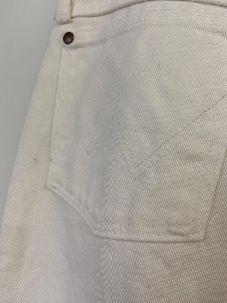 Op de afbeelding: Een close-up van een wit paar jeans met een achterzak. De zak heeft een gestikt ontwerp en een kleine knoop.