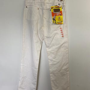 Op de afbeelding: Witte Wrangler jeans met een geel en rood label met de tekst "Cowboy Cut" en "Wrangler" hangend aan een hanger.