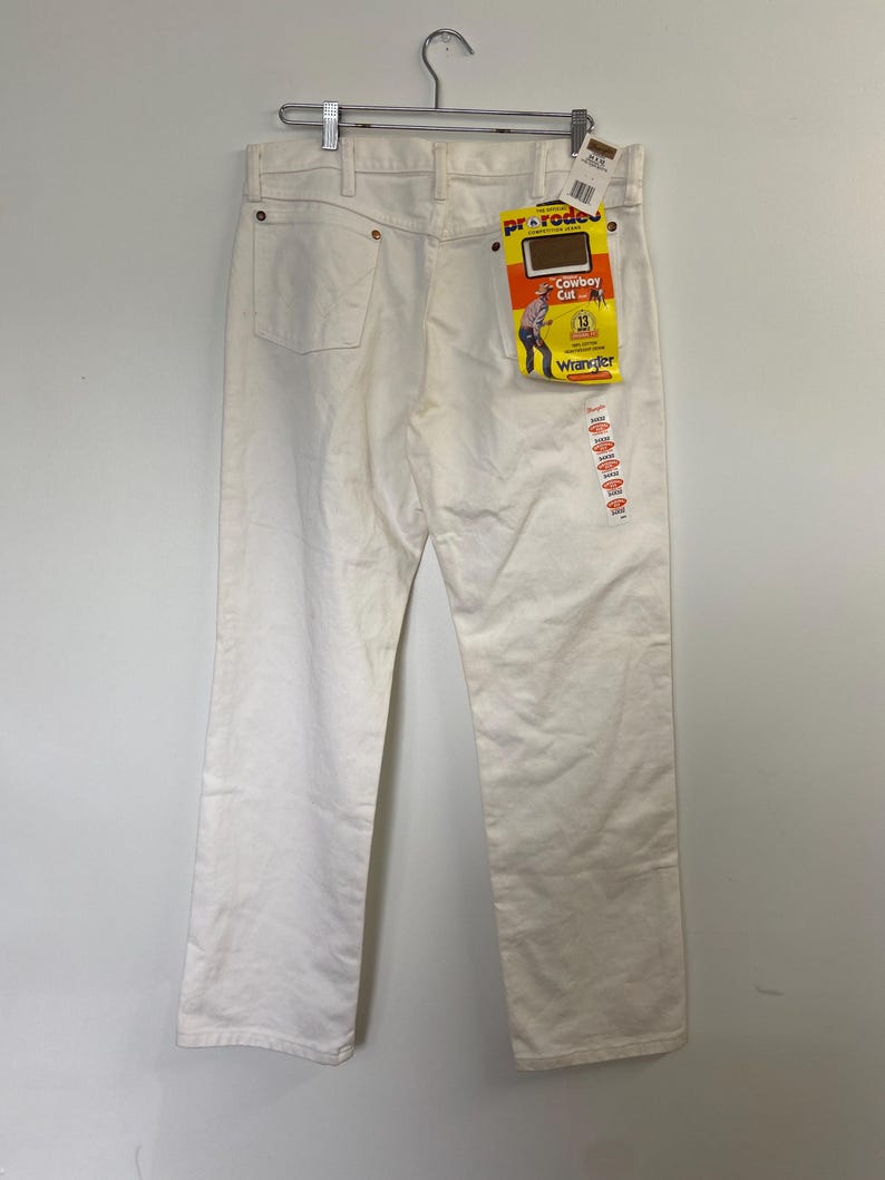 Op de afbeelding: Witte Wrangler jeans met een geel en rood label met de tekst "Cowboy Cut" en "Wrangler" hangend aan een hanger.