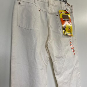 Op de afbeelding: Een witte spijkerbroek met een Wrangler ProRodeo-label eraan bevestigd. Het label toont een cowboy die een paard berijdt en de tekst "ProRodeo Competition Jeans" en "Wrangler".