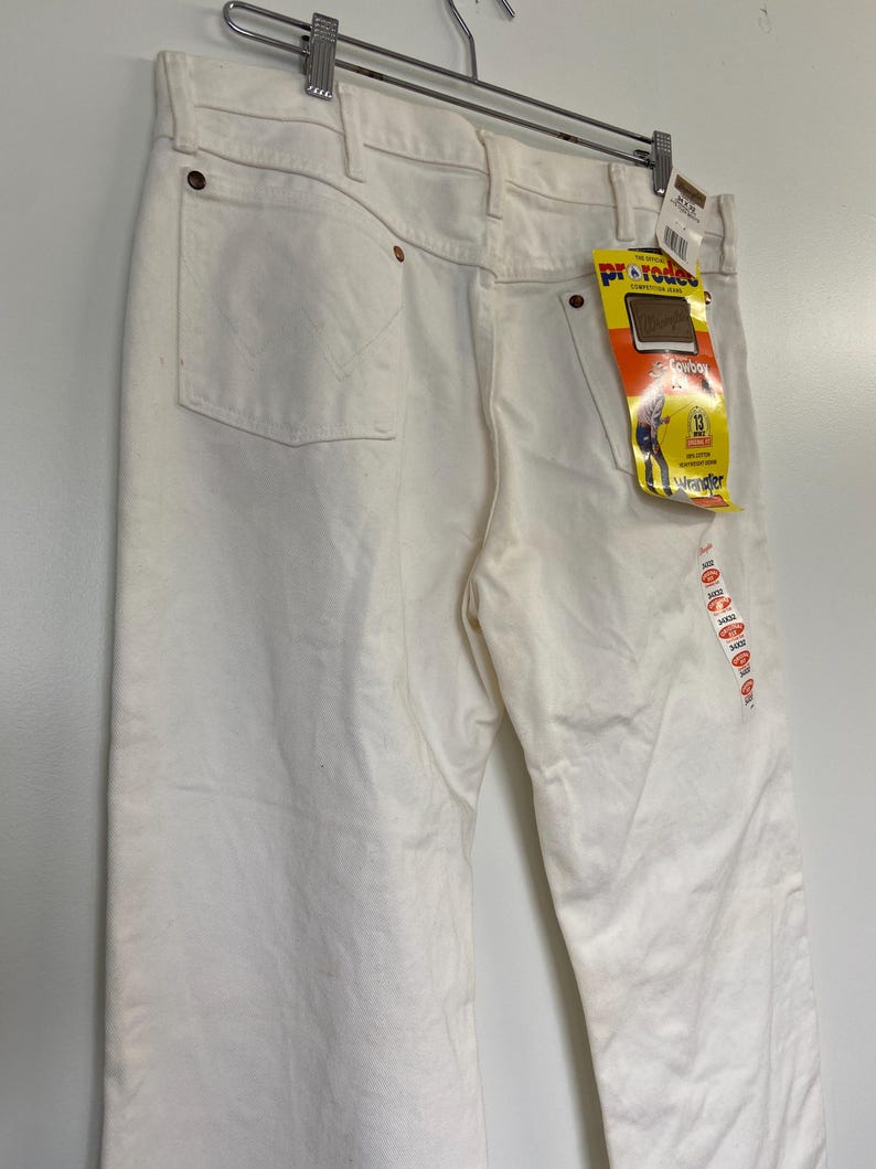 Op de afbeelding: Een witte spijkerbroek met een Wrangler ProRodeo-label eraan bevestigd. Het label toont een cowboy die een paard berijdt en de tekst "ProRodeo Competition Jeans" en "Wrangler".
