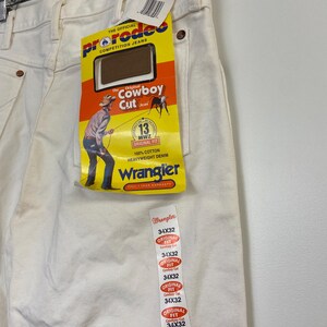 Op de afbeelding: Een witte spijkerbroek met een Wrangler-label eraan bevestigd. Het label heeft de tekst "Original Fit Cowboy Cut" en "34X32" erop gedrukt.