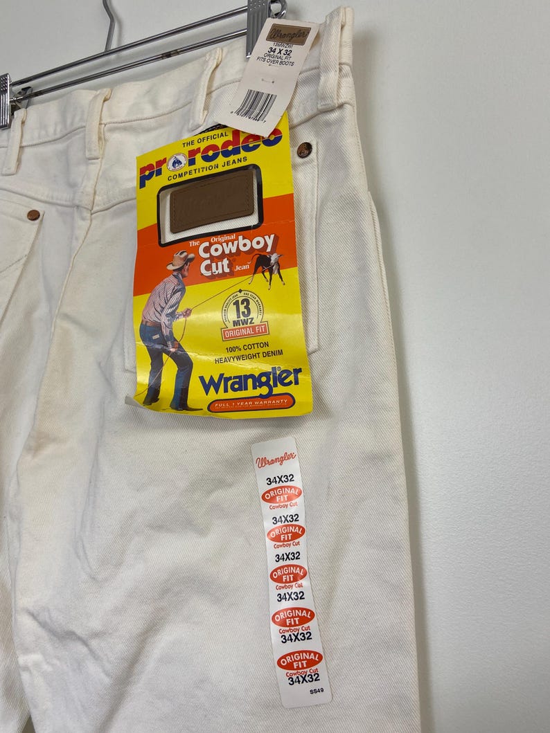 Op de afbeelding: Een witte spijkerbroek met een Wrangler-label eraan bevestigd. Het label heeft de tekst "Original Fit Cowboy Cut" en "34X32" erop gedrukt.