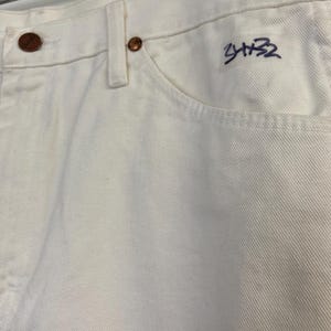 Op de afbeelding: Witte spijkerbroek met een paarse handtekening die "3432" op de rechterzak staat.